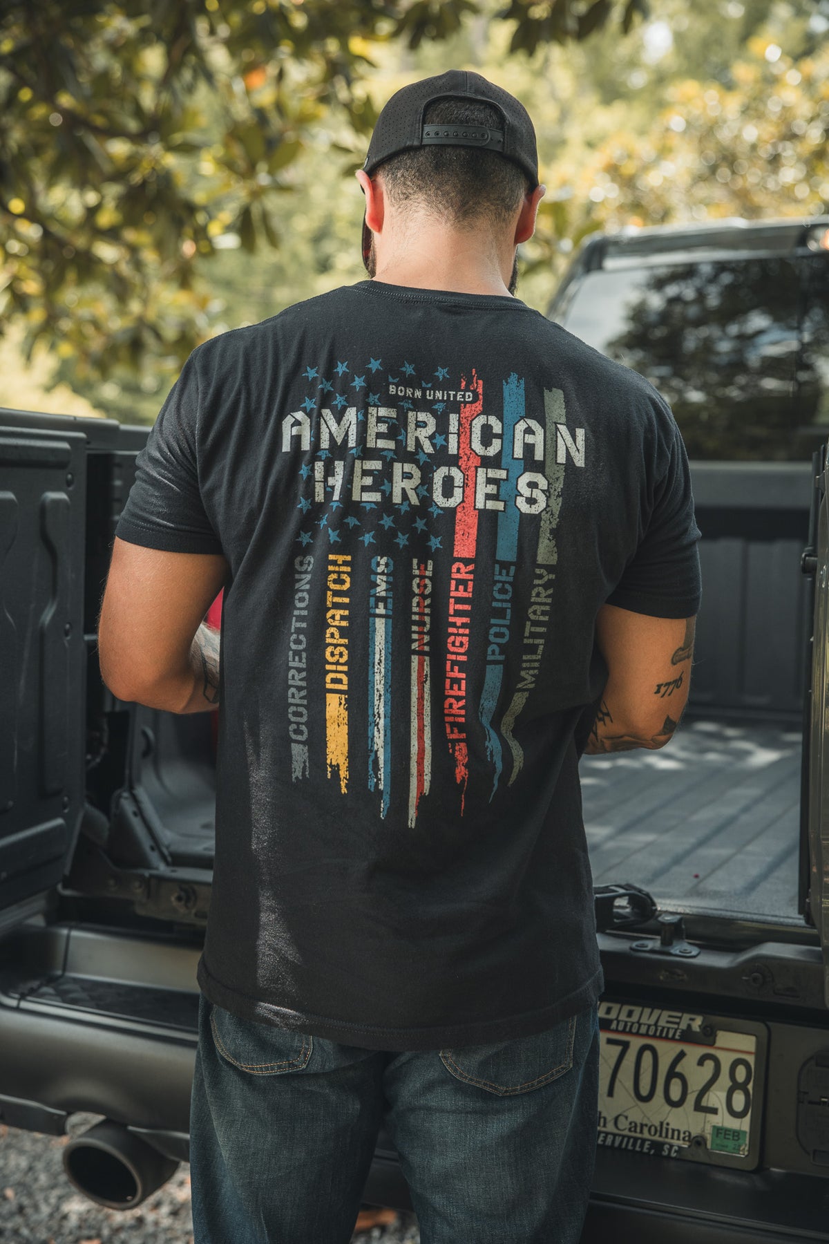 A man stands at the back of a pickup truck, wearing a black cap and an American Heroes Tee by Born United Private Label, featuring colorful stripes and words like Corrections, Dispatch, Firefighter, Police, and Military.