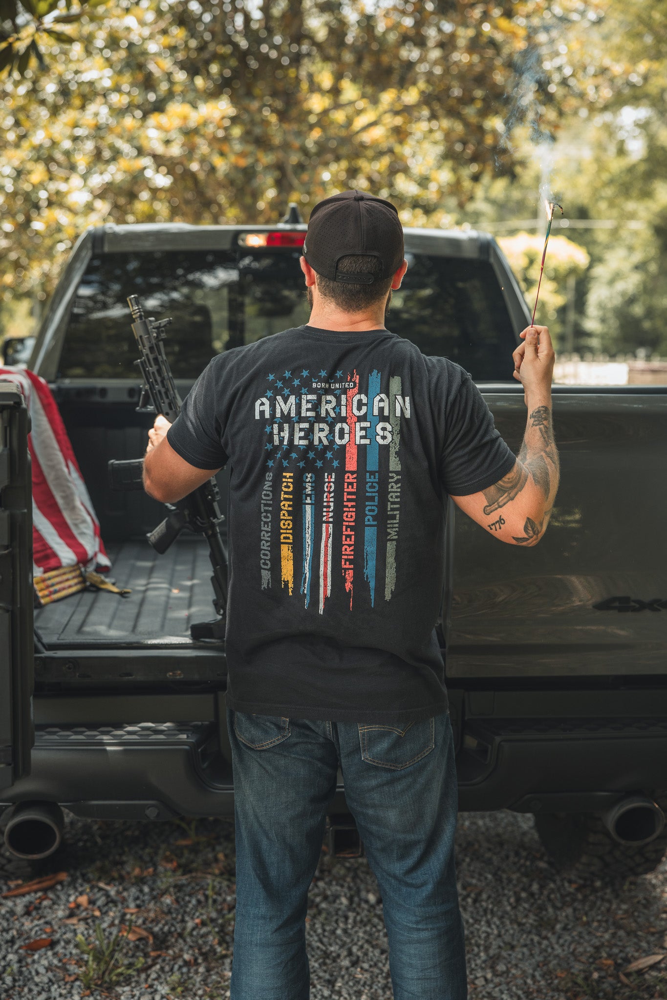 A man stands in a pickup truck holding a rifle and sparkler, wearing the Born United Private Label American Heroes Tee. The shirt features stripes and words like Honor and Sacrifice. An American flag is draped nearby.
