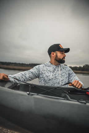A bearded man in a black cap and Mucker Bucket Performance Hoodie - Ghost Camo sits in a kayak on calm water, looking left, with an overcast sky and shoreline visible in the background.