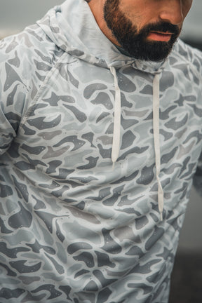A man with a dark beard models the Mucker Bucket Performance Hoodie - Ghost Camo, featuring light gray and white camouflage with white drawstrings. The cropped image shows his upper torso and part of his face.