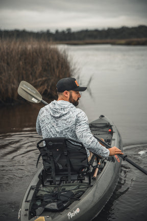 A man wearing a Mucker Bucket Performance Hoodie - Ghost Camo and a black cap paddles his kayak down a calm, overcast river bordered by reeds and grassy marshland.
