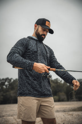 A man wearing the Mucker Bucket Performance Hoodie - Stealth Camo, a black hat, and beige shorts is outdoors with a fishing rod, adjusting the line against a backdrop of trees and a cloudy sky.