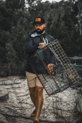 A man wears the Mucker Bucket Performance Hoodie - Stealth Camo, a dark jacket, and tan shorts as he carries a metal cage trap outdoors among trees and rough terrain.