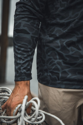 A person wearing the Mucker Bucket Performance Hoodie - Stealth Camo and beige pants holds several coiled ropes in their left hand near a wooden structure outdoors, with the focus on their torso and hand.