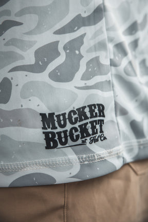 Close-up of the Mucker Bucket Performance Hoodie in Ghost Camo, featuring a light gray and white camouflage pattern with the brand printed in black, draped over tan pants.