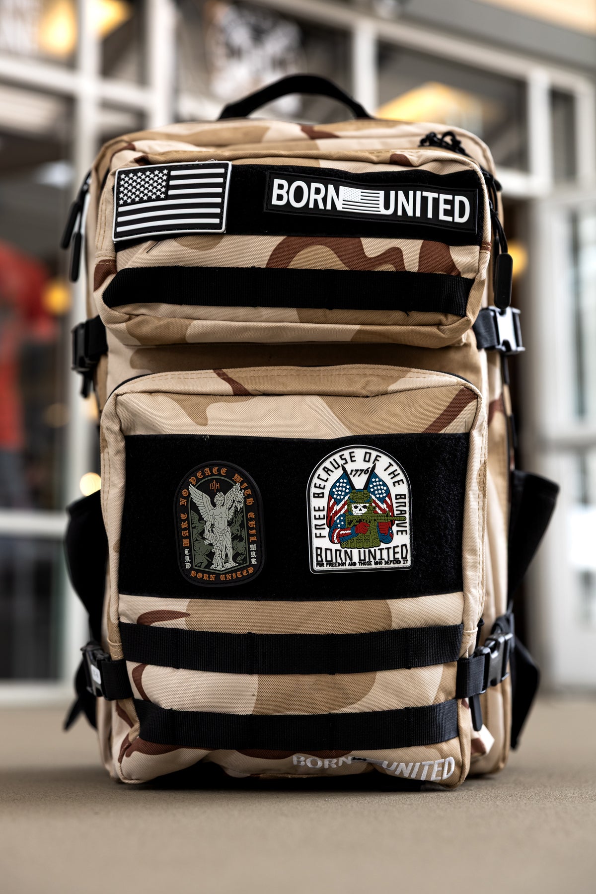 The OIF Camo Premium Tac-Pac - 45L by Born United Private Label is a tan and brown camouflage tactical backpack with MOLLE loops and multiple patches, including an American flag and BORN UNITED, shown upright indoors with glass windows behind.