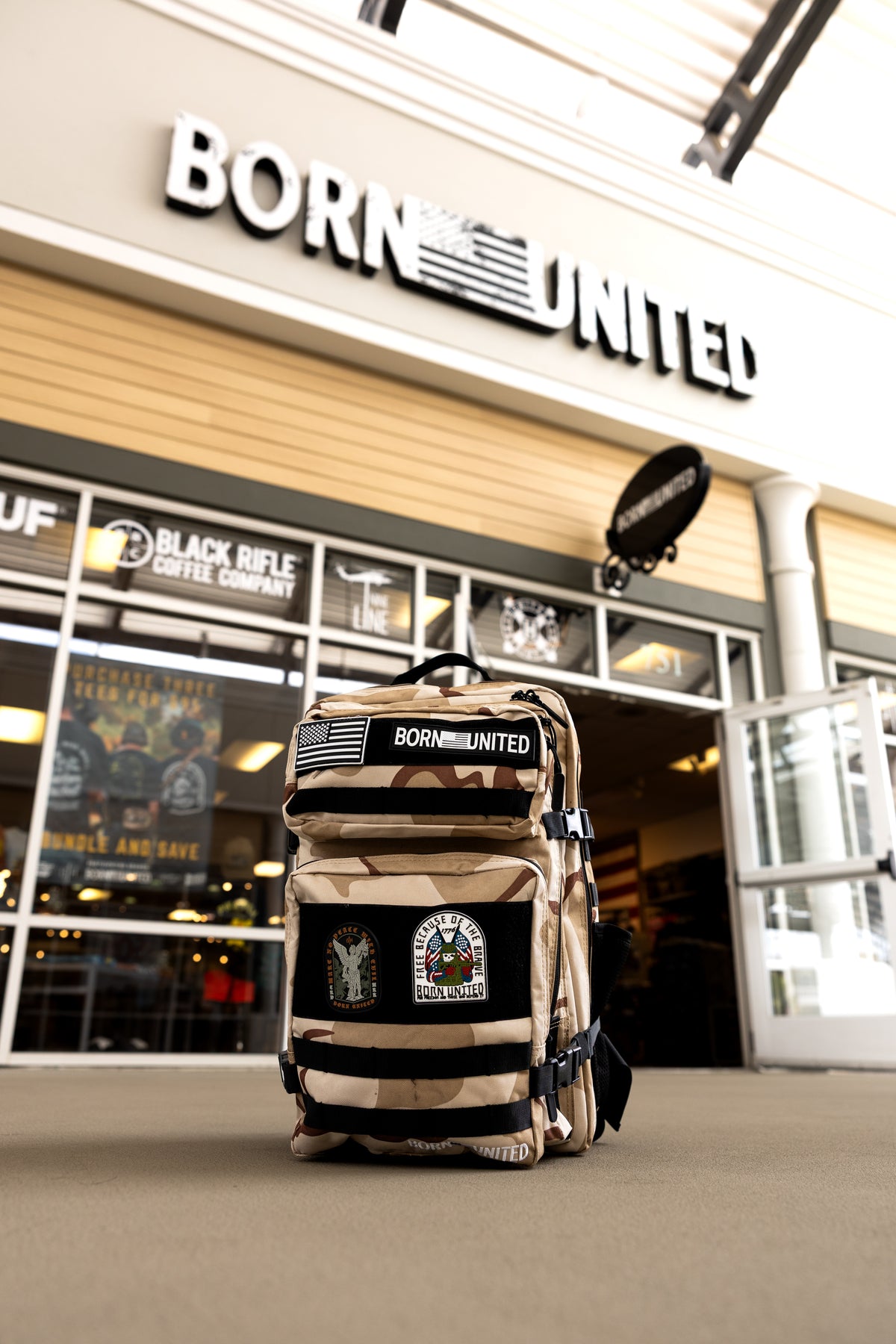 The OIF Camo Premium Tac-Pac - 45L by Born United Private Label, with MOLLE loops and BORN UNITED patches, is displayed on the ground in front of a Born United store and a Black Rifle Coffee Company sign.