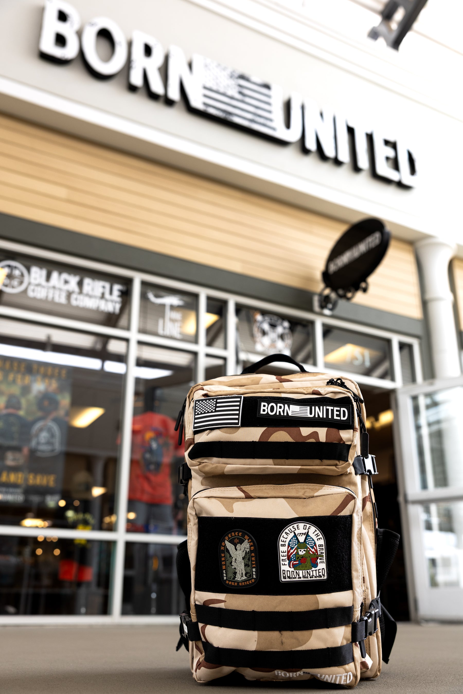 The OIF Camo Premium Tac-Pac - 45L by Born United Private Label, featuring tan camo and MOLLE loops, is displayed on the ground in front of a BORN UNITED store with its sign and entrance visible in the background.