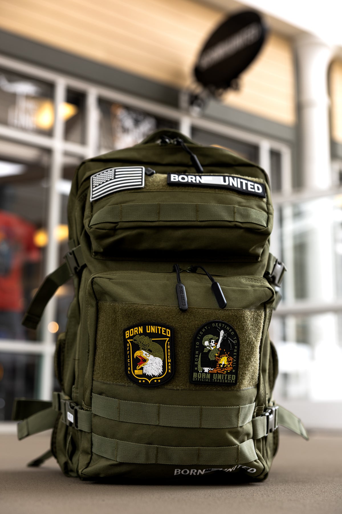 The Born United Private Label OD Green Premium Tac-Pac - 45L, decorated with American flag, BORN UNITED, eagle, and skull patches, rests on an outdoor table in front of a building with large windows.