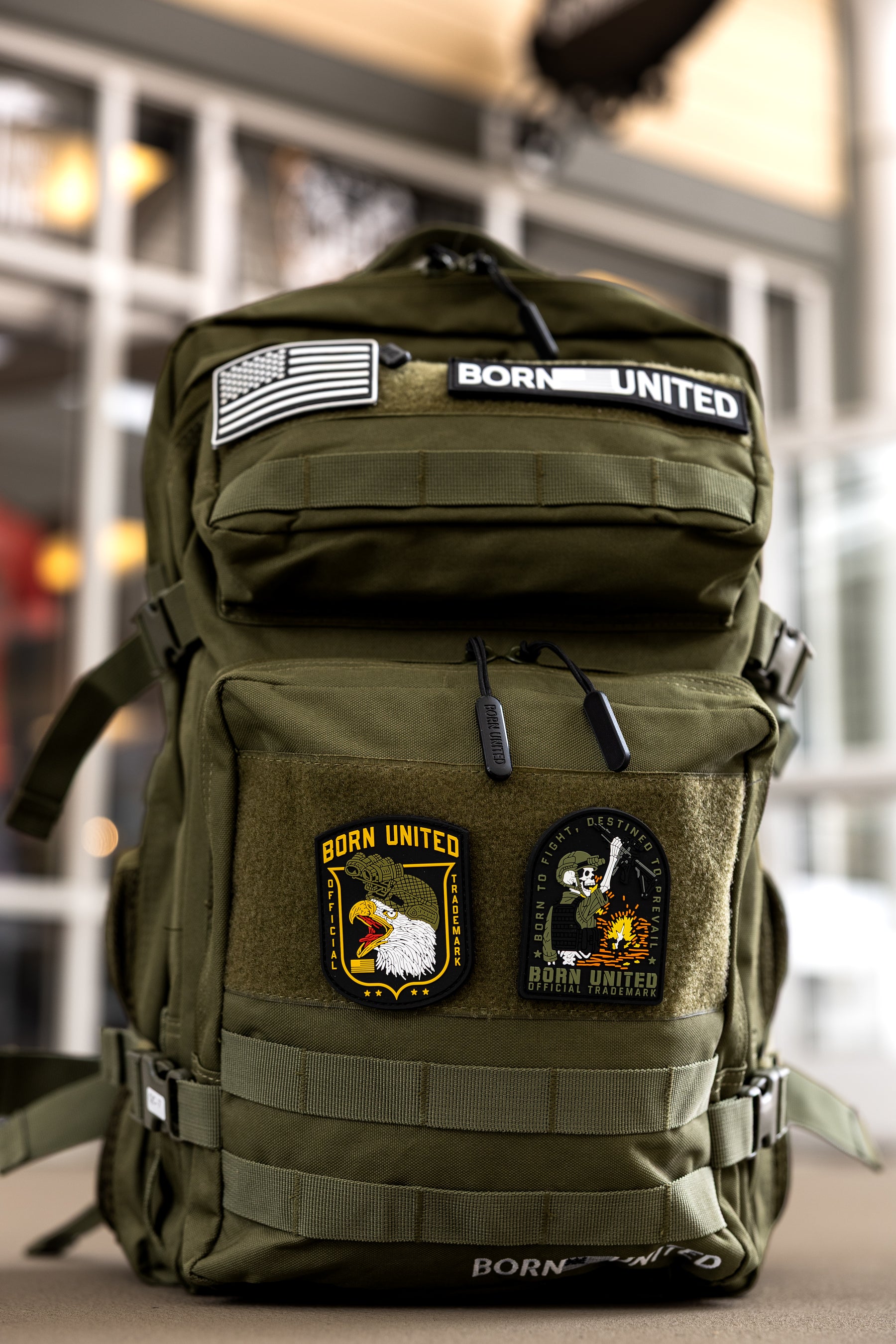 The Born United Private Label OD Green Premium Tac-Pac 45L, featuring American flag, BORN UNITED, eagle, and skull patches plus TSA-approved features, sits on a table indoors with a blurred background for focus.