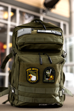 The Born United Private Label OD Green Premium Tac-Pac 45L, featuring American flag, BORN UNITED, eagle, and skull patches plus TSA-approved features, sits on a table indoors with a blurred background for focus.