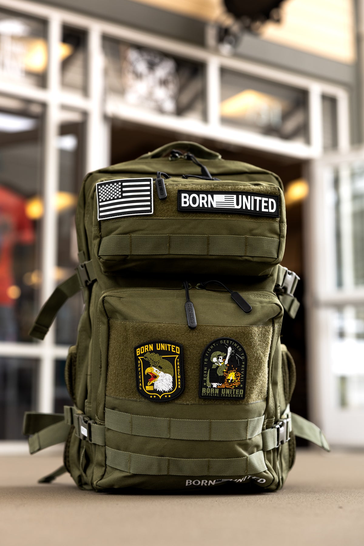 The OD Green Premium Tac-Pac - 45L by Born United Private Label, decorated with patches like an American flag, BORN UNITED, an eagle, and a rocket, stands upright on the floor in front of a windowed building.