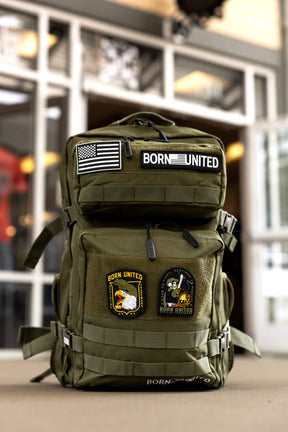 The OD Green Premium Tac-Pac - 45L by Born United Private Label, decorated with patches like an American flag, BORN UNITED, an eagle, and a rocket, stands upright on the floor in front of a windowed building.