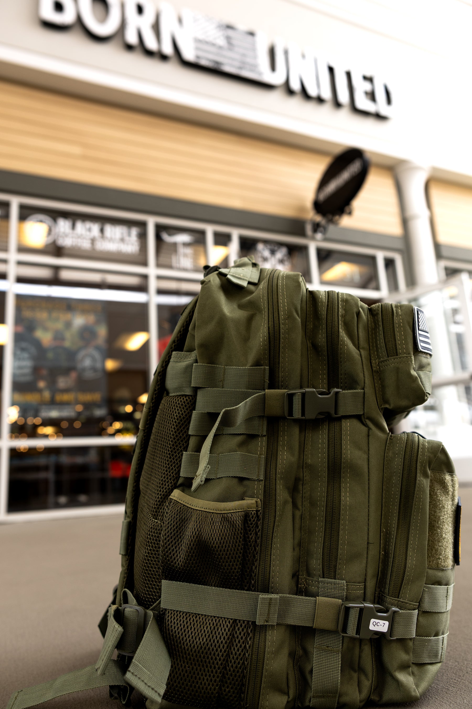 The OD Green Premium Tac-Pac - 45L by Born United Private Label sits on the ground outside a store with large glass windows and a blurry Born United sign above the entrance.