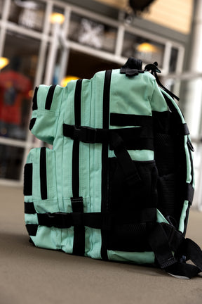 The Mint Condition Tac-Pac - 25L by Born United Private Label, featuring mint green and black colors, multiple pockets, organized storage, and a modular patch panel, stands upright on a brown surface with windows and indoor lighting in the background.