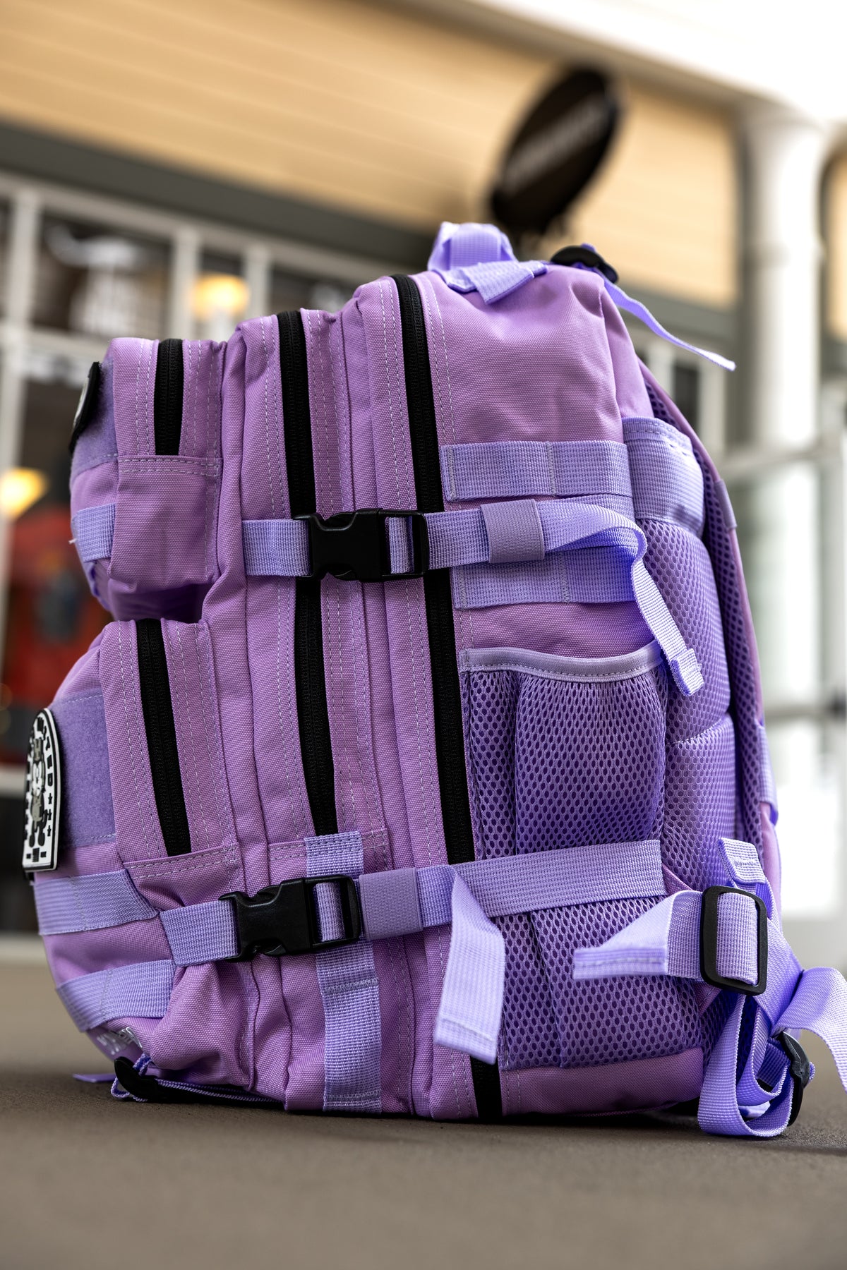 The Imperial Purple Tac-Pac - 25L by Born United Private Label, featuring multiple black zippers, mesh pockets, and adjustable straps, rests outdoors on a flat surface with a blurred building in the background.