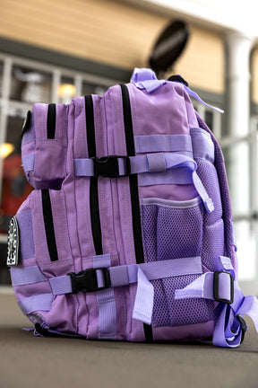 The Imperial Purple Tac-Pac - 25L by Born United Private Label, featuring multiple black zippers, mesh pockets, and adjustable straps, rests outdoors on a flat surface with a blurred building in the background.