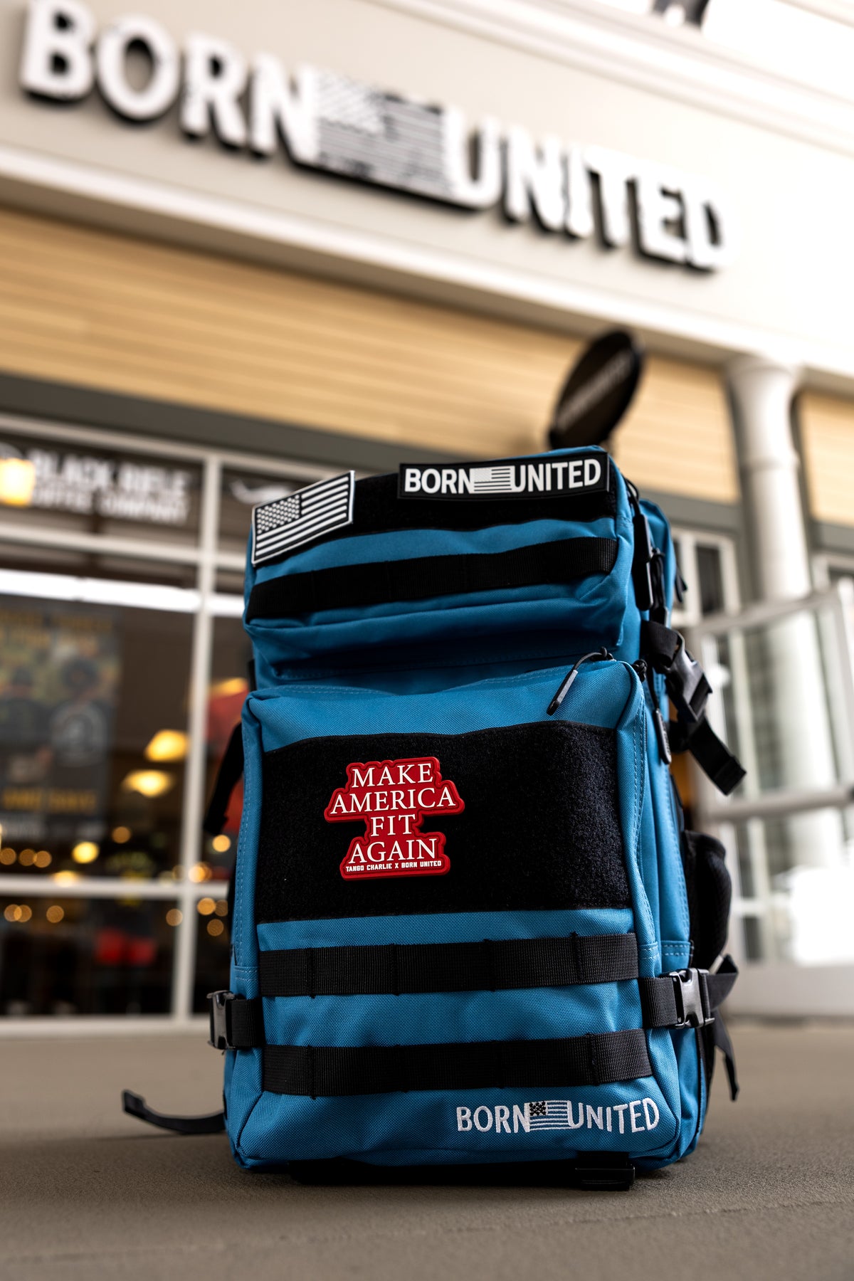 A Peacock Blue Limited Ed. Tac-Pac - 45L by Born United Private Label, featuring a red Make America Fit Again patch, sits on the ground before a store with a blurred Born United sign above the entrance.