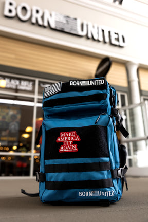 A Peacock Blue Limited Ed. Tac-Pac - 45L by Born United Private Label, featuring a red Make America Fit Again patch, sits on the ground before a store with a blurred Born United sign above the entrance.