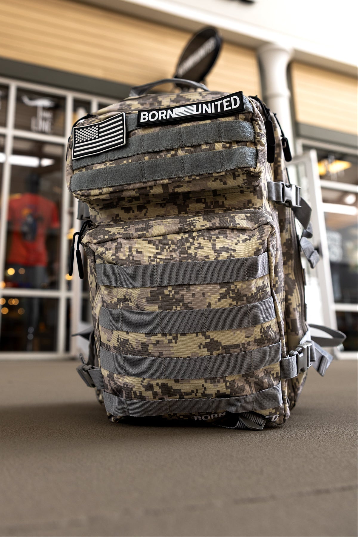 The Born United Private Label ACU Tac-Pac - 45L, featuring an American flag patch and BORN UNITED patch, rests outside a store with a BORN UNITED sign above the entrance.