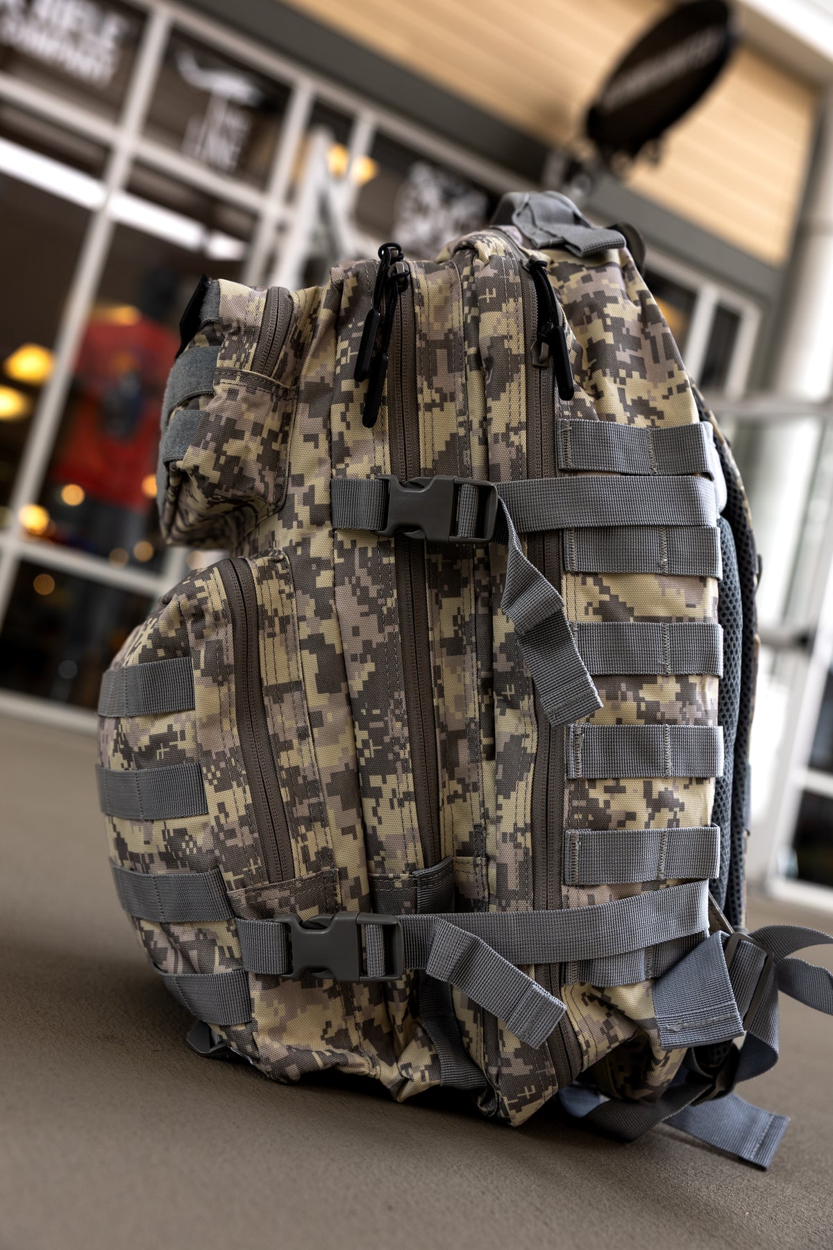 A Born United Private Label ACU Tac-Pac - 45L tactical backpack with multiple zippers, straps, and buckles rests on the ground in front of a building with glass doors and windows.