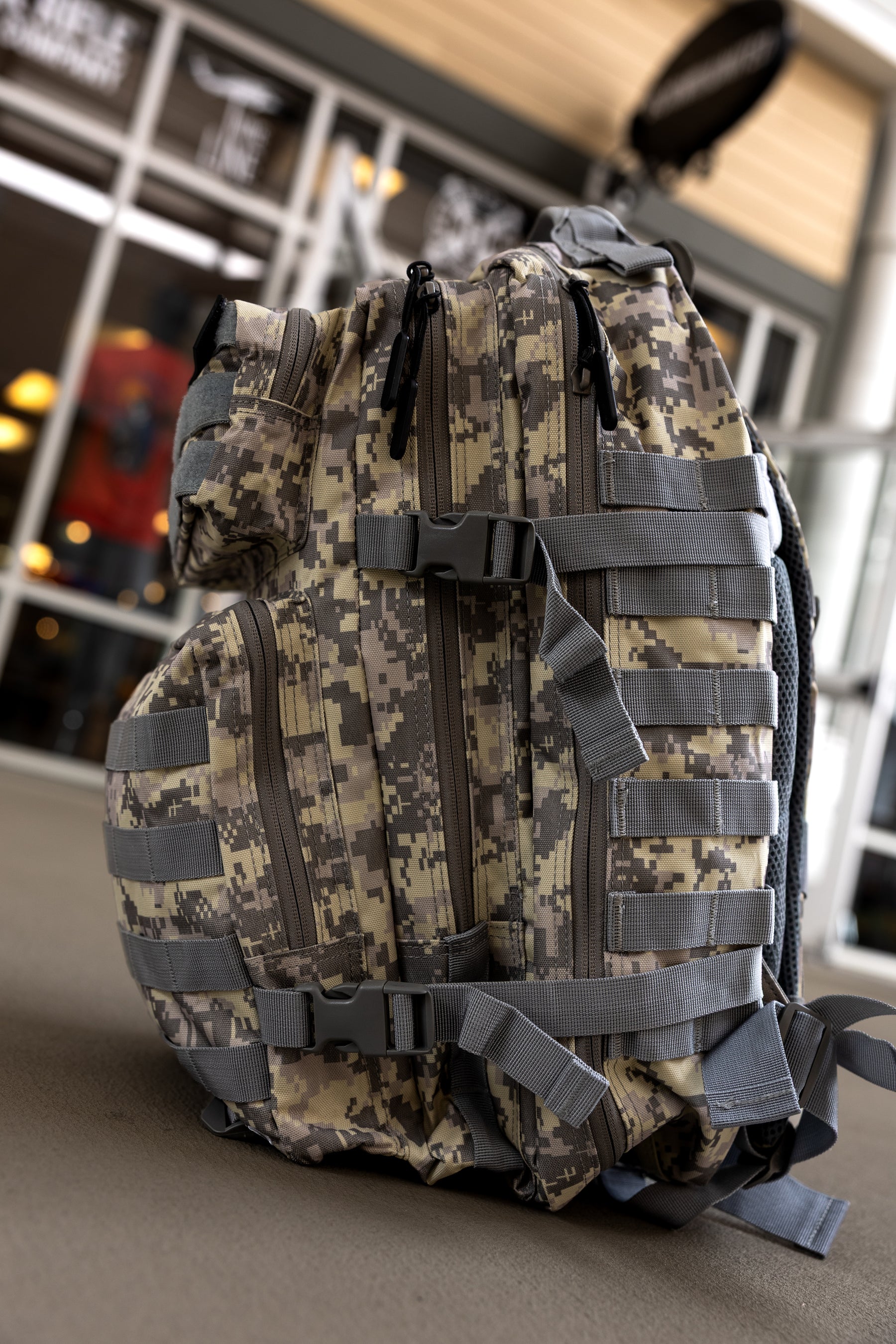 A Born United Private Label ACU Tac-Pac - 45L tactical backpack with multiple zippers, straps, and buckles rests on the ground in front of a building with glass doors and windows.