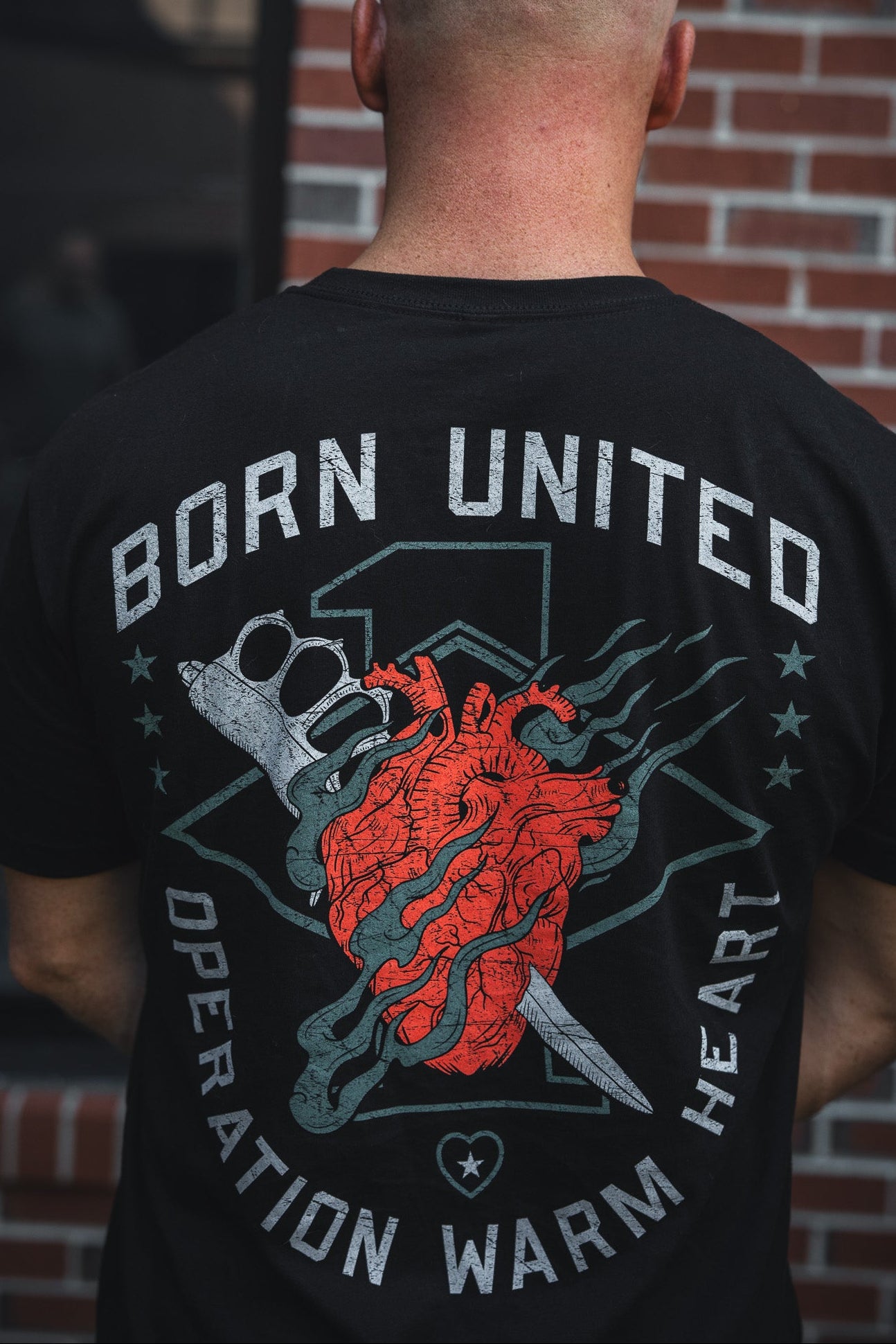 A person with a shaved head wears the Born United Private Label’s Operation Warm Heart - Black tee, featuring a red heart, brass knuckles, and knife. “BORN UNITED” tops the shirt; “OPERATION WARM HEART” is below—supporting America’s heroes.