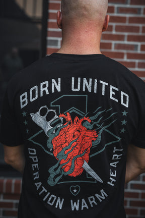 A person with a shaved head wears the Born United Private Label’s Operation Warm Heart - Black tee, featuring a red heart, brass knuckles, and knife. “BORN UNITED” tops the shirt; “OPERATION WARM HEART” is below—supporting America’s heroes.