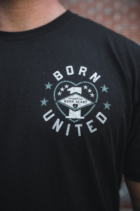 Close-up of a person wearing the Born United Private Label “Operation Warm Heart - Black” limited edition tee, featuring “BORN UNITED” and a circular emblem on the front. A blurred brick wall background honors support for America’s heroes.