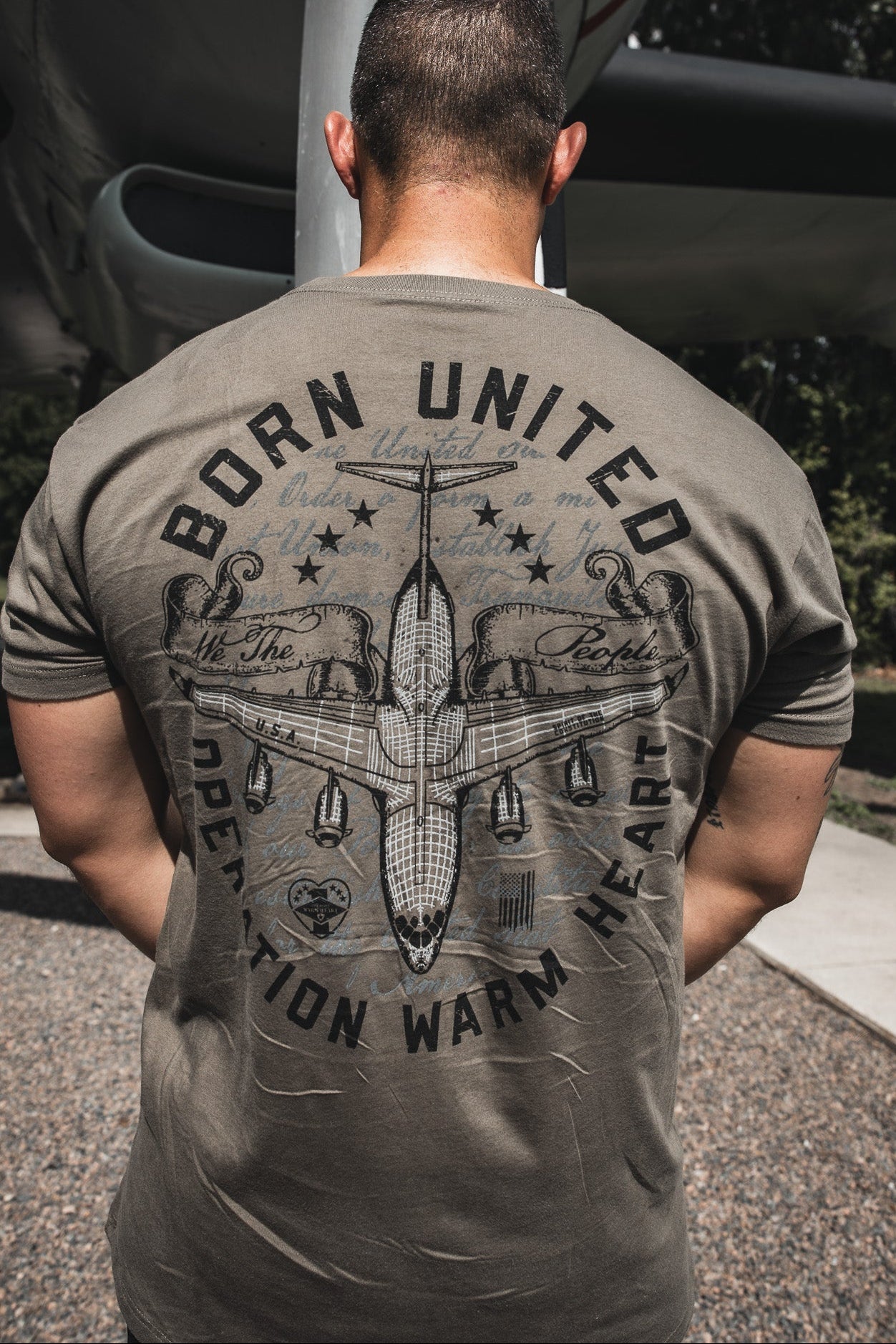 At Joint Base Charleston, a man faces away from the camera wearing the Born United Private Label "Operation Warm Heart- Olive" T-shirt, featuring military-inspired graphics and an aircraft in the background.