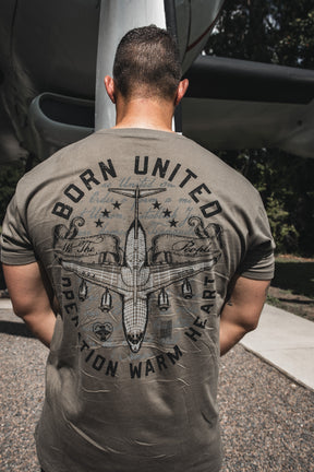 At Joint Base Charleston, a man faces away from the camera wearing the Born United Private Label "Operation Warm Heart- Olive" T-shirt, featuring military-inspired graphics and an aircraft in the background.