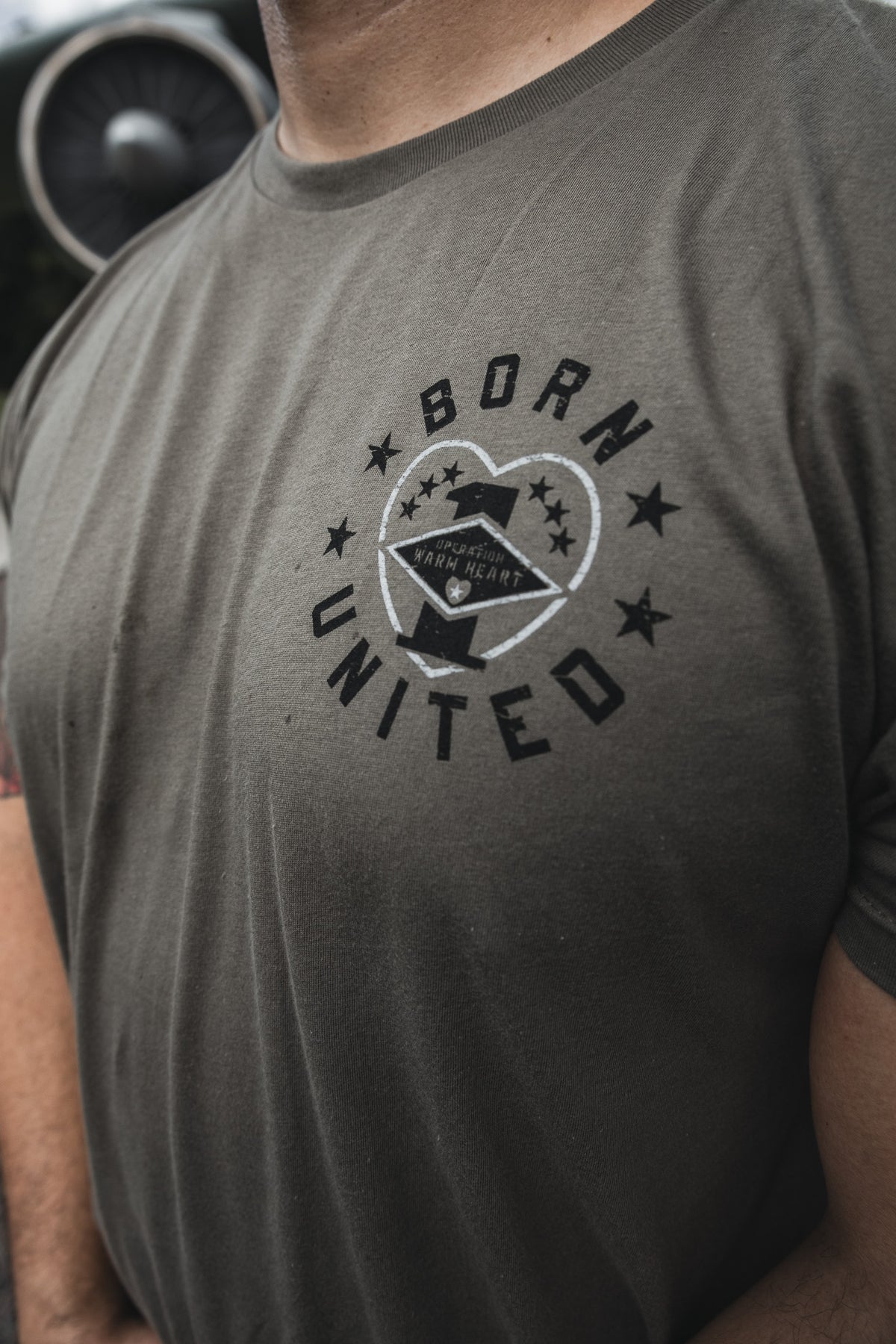 A person wears the Operation Warm Heart-Olive t-shirt by Born United Private Label, featuring a circular "BORN UNITED" logo with a star and eagle, standing outdoors at Joint Base Charleston with an aircraft engine blurred in the background.