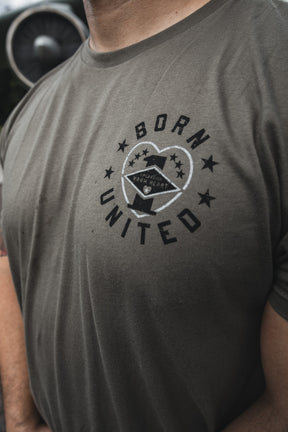 A person wears the Operation Warm Heart-Olive t-shirt by Born United Private Label, featuring a circular "BORN UNITED" logo with a star and eagle, standing outdoors at Joint Base Charleston with an aircraft engine blurred in the background.