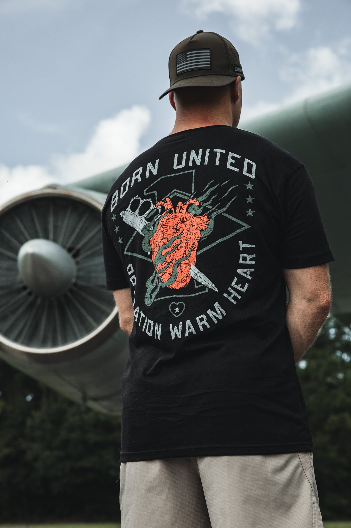 Wearing the Born United Private Label Operation Warm Heart - Black tee, featuring a bold anatomical heart graphic, a supporter stands outdoors by an airplane engine, honoring America’s heroes.