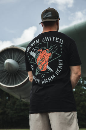 Wearing the Born United Private Label Operation Warm Heart - Black tee, featuring a bold anatomical heart graphic, a supporter stands outdoors by an airplane engine, honoring America’s heroes.