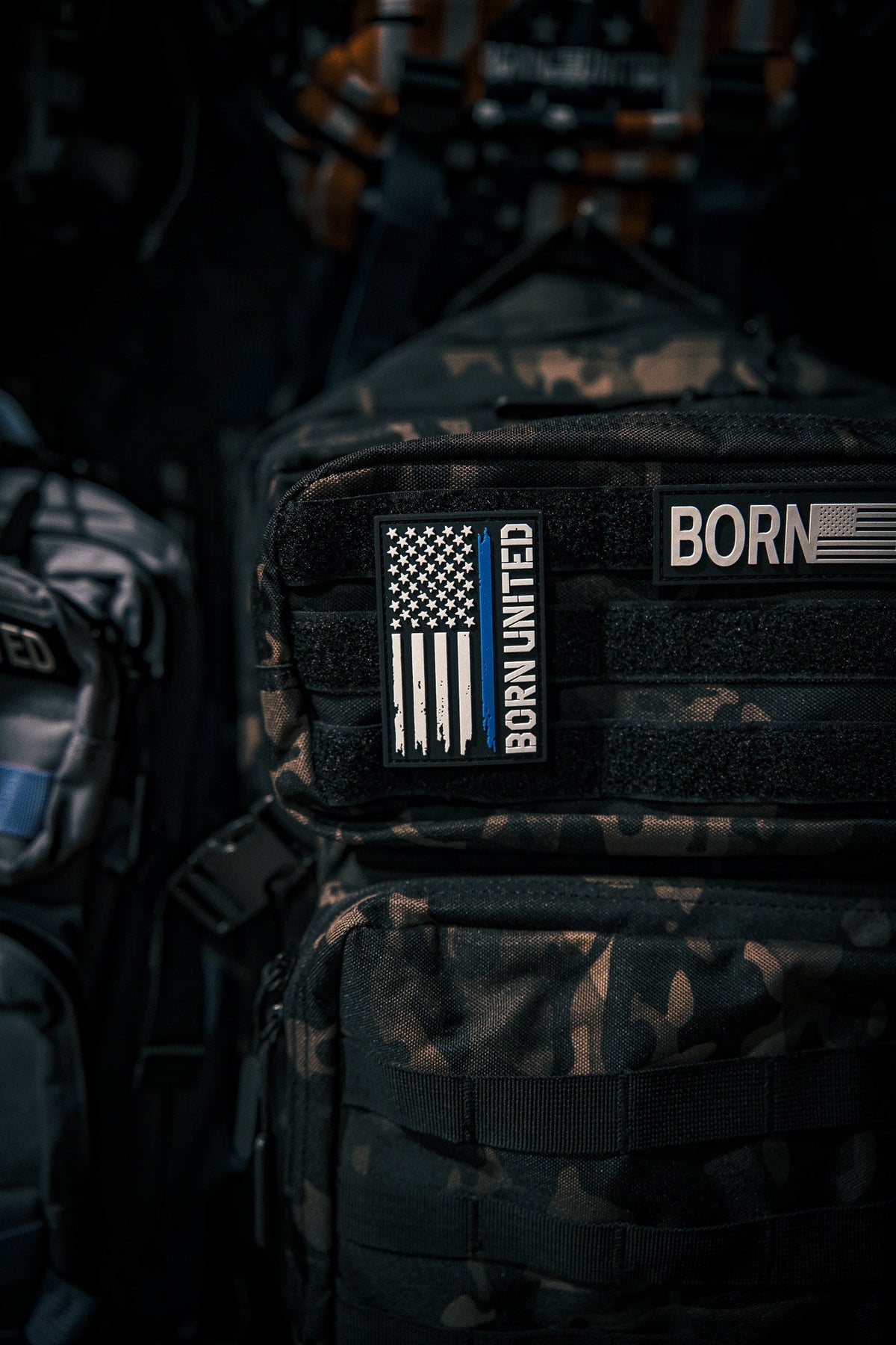 A camouflage backpack displays the Born United Private Label Blue Line Flag PVC Patch featuring a black-and-white American flag with a blue stripe. More backpacks and gear appear in the background.