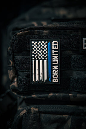 A Blue Line Flag PVC Patch by Born United Private Label, featuring a black, white, and blue American flag with a single blue stripe and BORN UNITED text, is attached to a black tactical backpack.