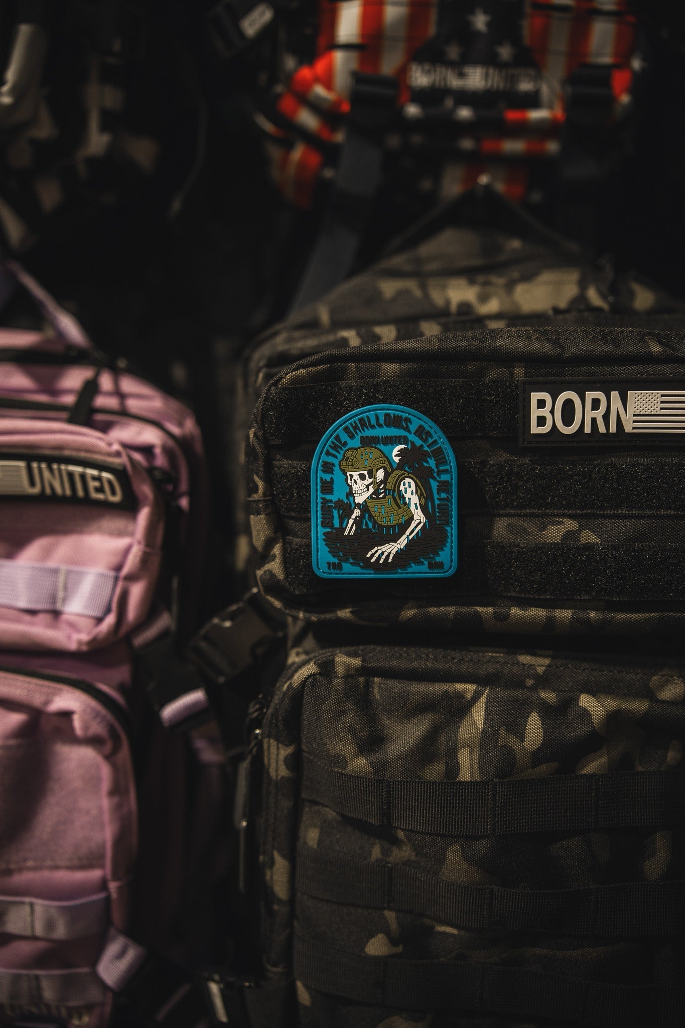 A close-up of a black camouflage backpack with The Shallows PVC Patch by Born United Private Label, showing a skeleton holding a rifle. Nearby, other black and pink backpacks feature visible BORN and UNITED labels and similar patches.