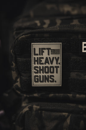 A close-up of a camouflage backpack featuring the Lift Heavy. Shoot Guns. - PVC Patch by Tango Charlie Apparel, displaying bold black lettering on a gray background.