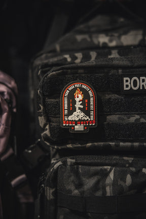 The Let The Darkness PVC Patch by Born United Private Label, featuring a flaming torch, red and white details, stars, and edge text, is attached to the front pocket of a black camo-patterned backpack.