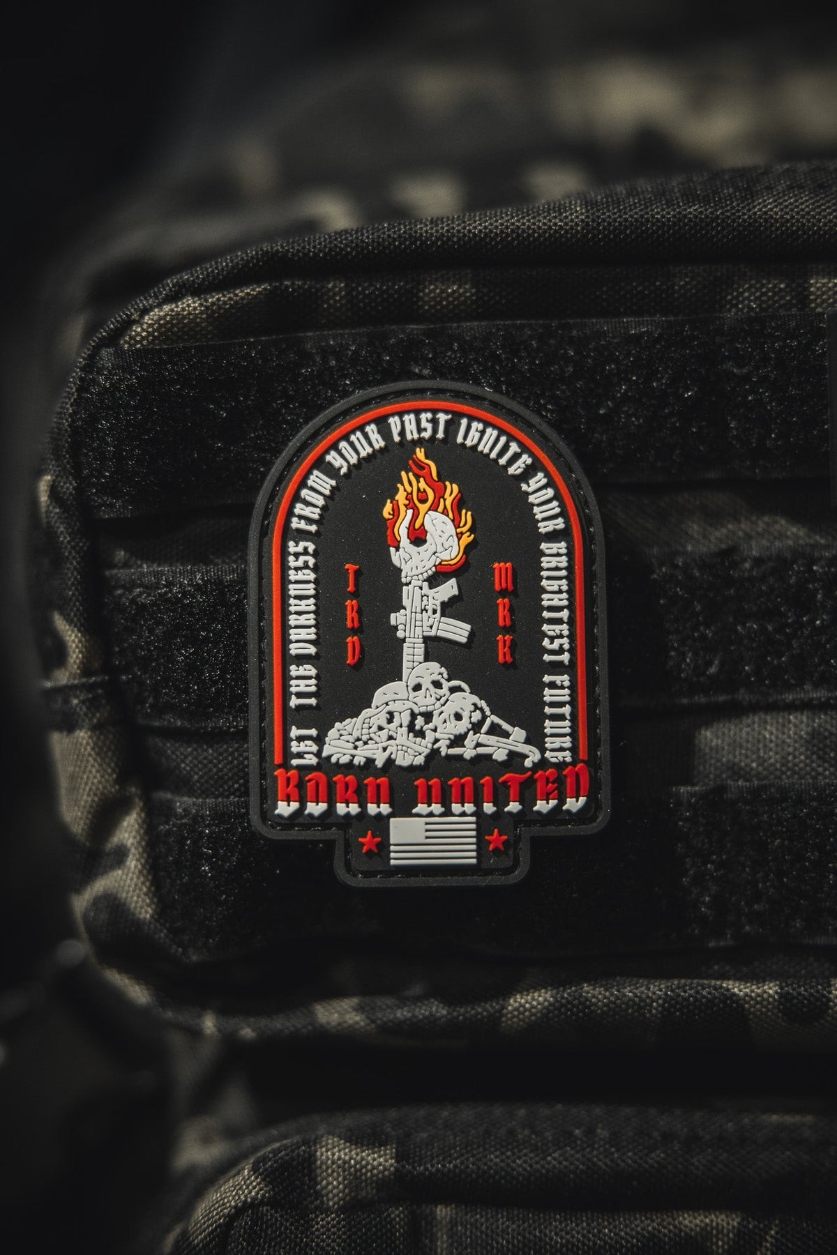 The Let The Darkness PVC Patch by Born United Private Label features a fiery torch above skulls and bones with BORN UNITED and phrases encircling the torch, all set on textured black fabric.