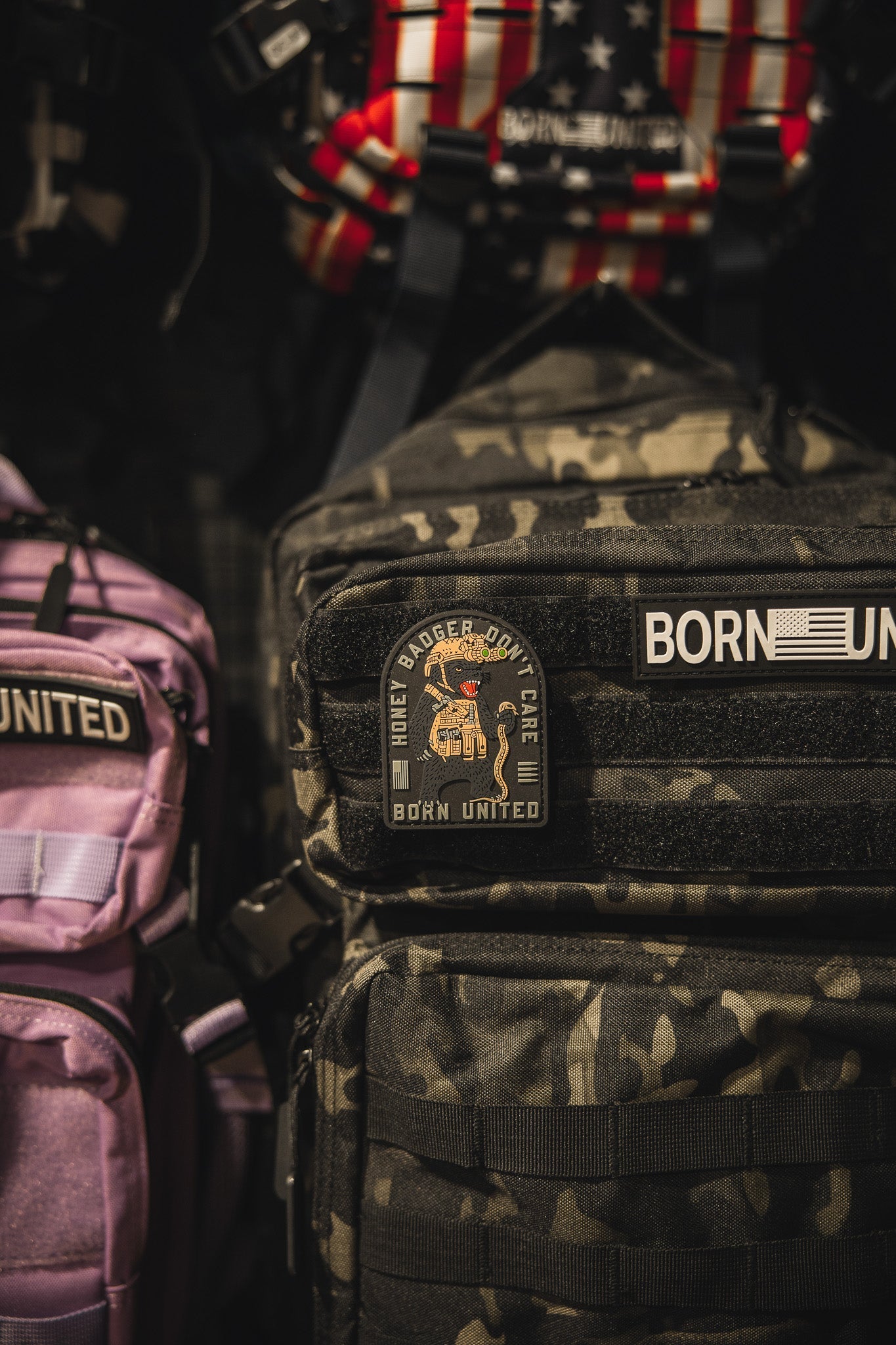 A close-up of camouflage and purple backpacks with patches, including the Honey Badgerin PVC Patch from Born United Private Label on the camo bag, featuring a cartoon badger and nearby US flag-themed patch.