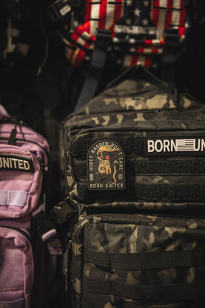 A close-up of camouflage and purple backpacks with patches, including the Honey Badgerin PVC Patch from Born United Private Label on the camo bag, featuring a cartoon badger and nearby US flag-themed patch.