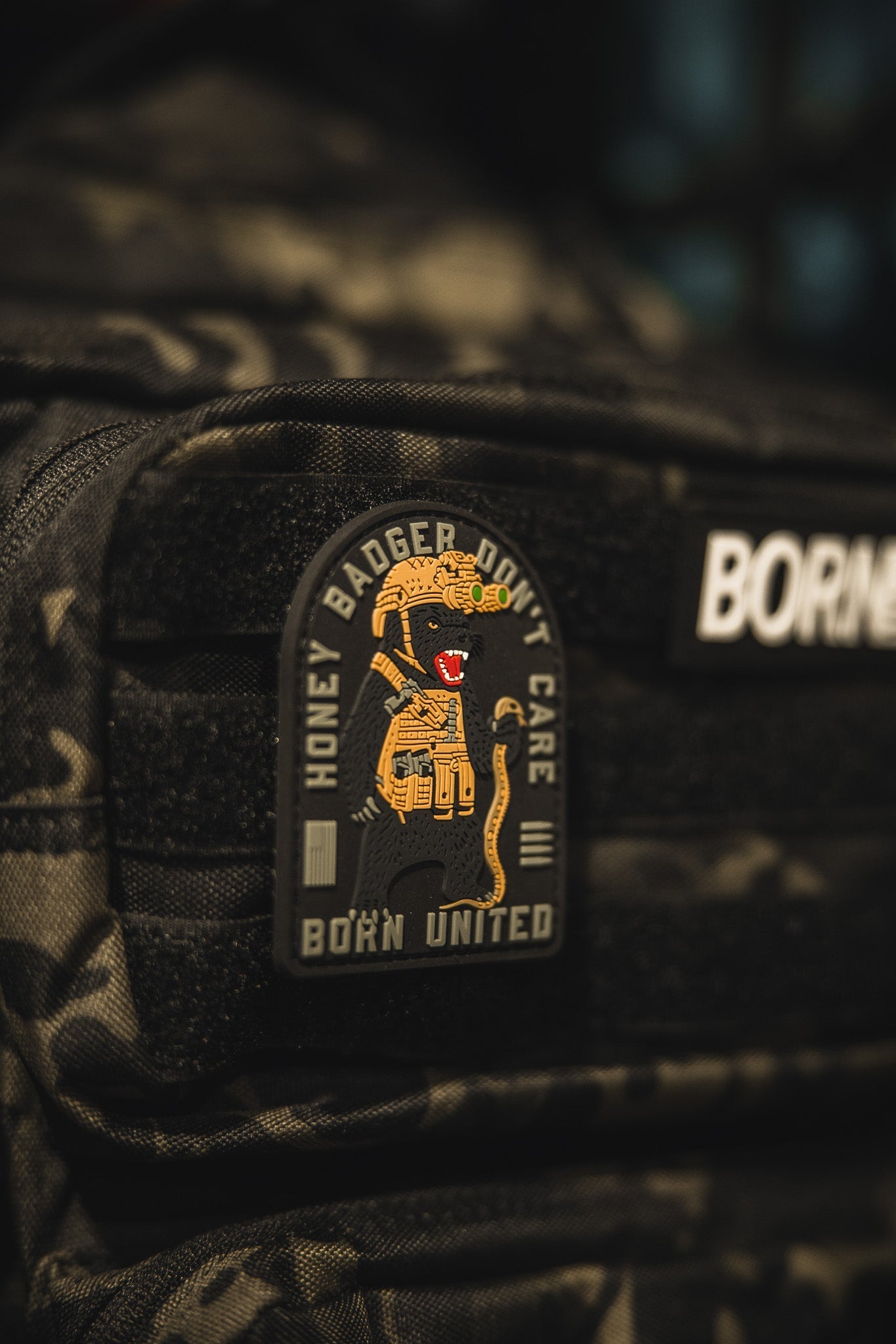 A close-up of a black tactical backpack with the Born United Private Label Honey Badgerin PVC Patch, featuring a cartoon honey badger holding a rifle and the text HONEY BADGER DONT CARE and BORN UNITED.