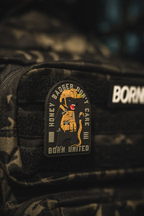 A close-up of a black tactical backpack with the Born United Private Label Honey Badgerin PVC Patch, featuring a cartoon honey badger holding a rifle and the text HONEY BADGER DONT CARE and BORN UNITED.