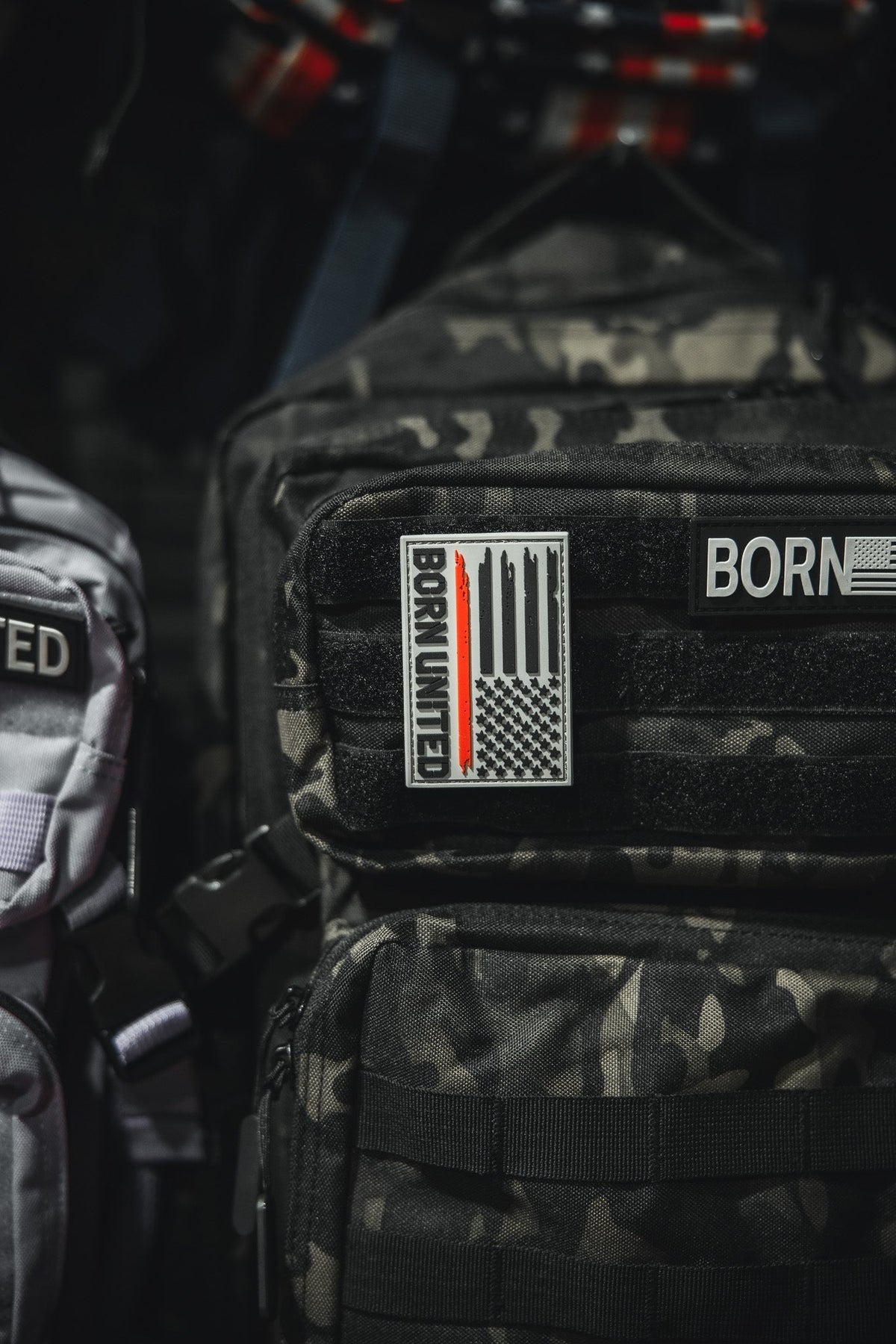 A black camouflage backpack displays a Red Line Flag PVC Patch by Born United Private Label with a stylized U.S. flag and BORN UNITED text. Above it is another partial BORN patch. A light gray bag appears partially on the left.