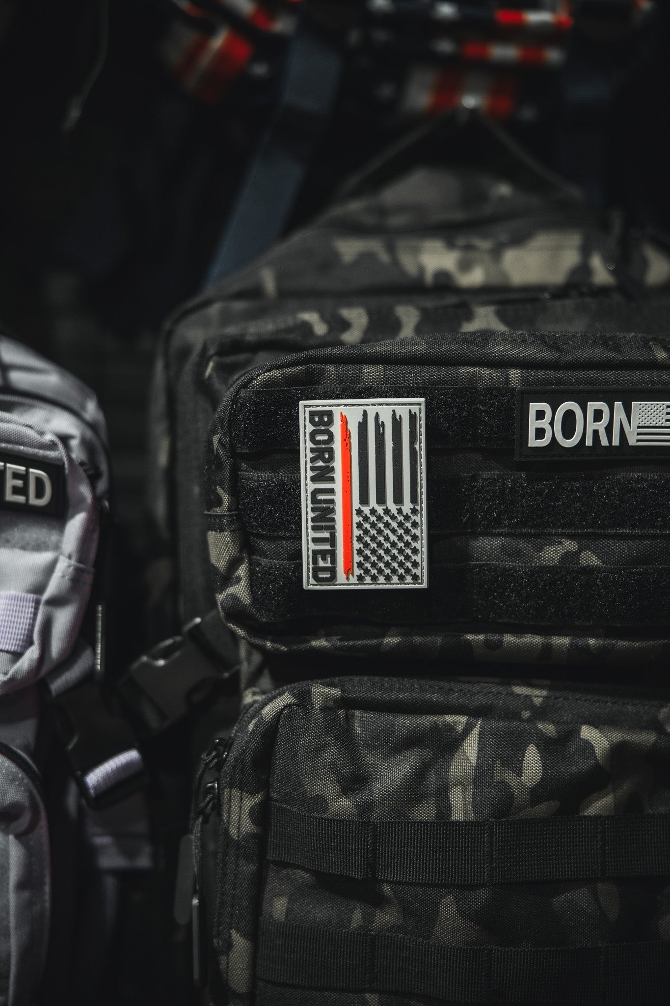 A black camouflage backpack displays a Red Line Flag PVC Patch by Born United Private Label with a stylized U.S. flag and BORN UNITED text. Above it is another partial BORN patch. A light gray bag appears partially on the left.