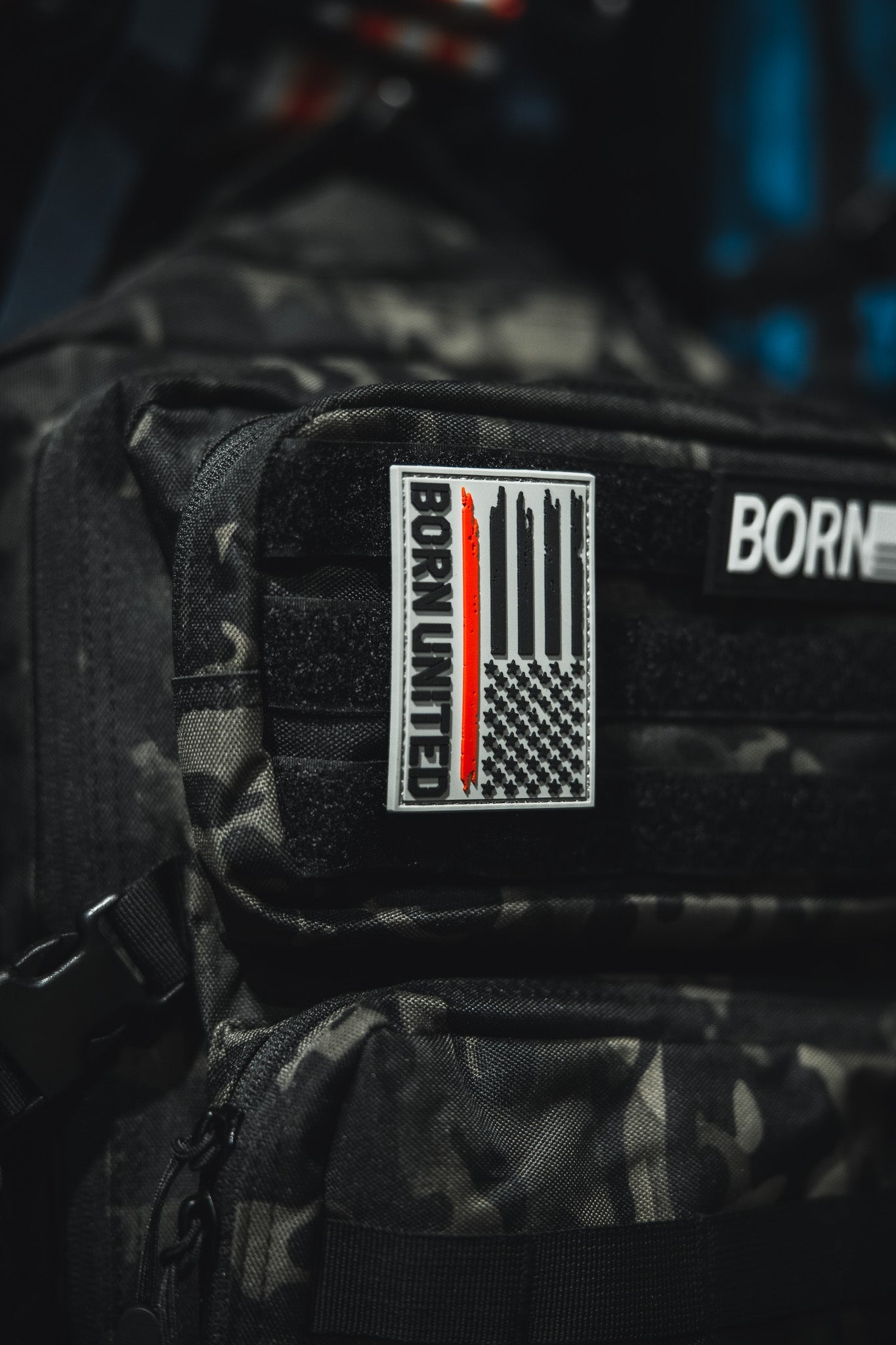 A close-up of a black camo backpack displays the Red Line Flag PVC Patch by Born United Private Label, featuring a grayscale American flag with a single red stripe and bold BORN UNITED text.