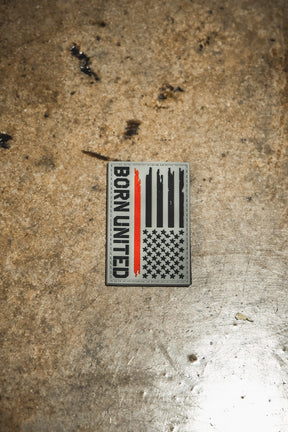 The Red Line Flag PVC Patch by Born United Private Label is a rectangular patch with a stylized American flag, featuring one red stripe and “BORN UNITED” vertically on the left, displayed on a worn, brownish metal surface.