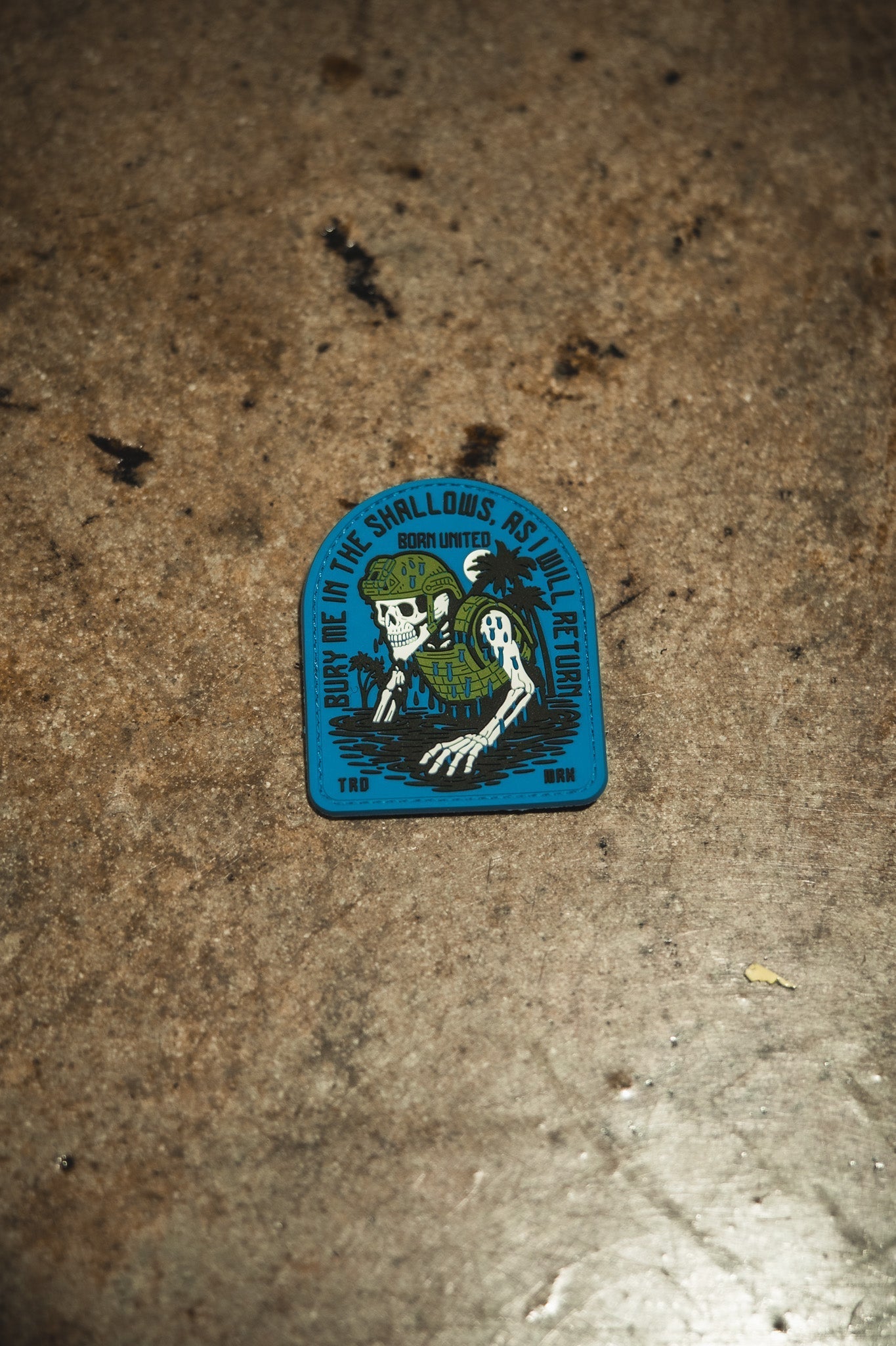 A rugged PVC patch by Born United Private Label shows a skeleton with seaweed and helmet, gripping a trident, plus the text: “Bury me in the shallows, but I’ll return. Born united. TRD. BNM.” Product: The Shallows PVC Patch.